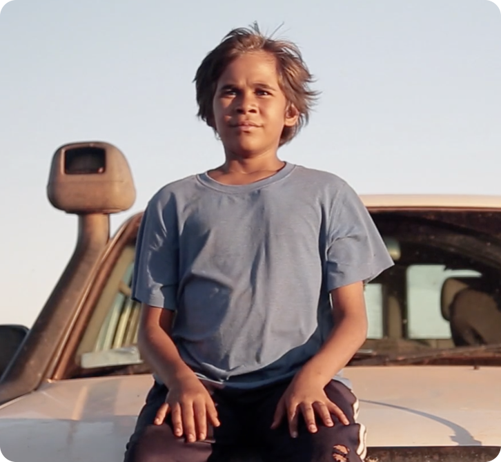 Young boy sits on the bonnet of a car looking out into the sunset.